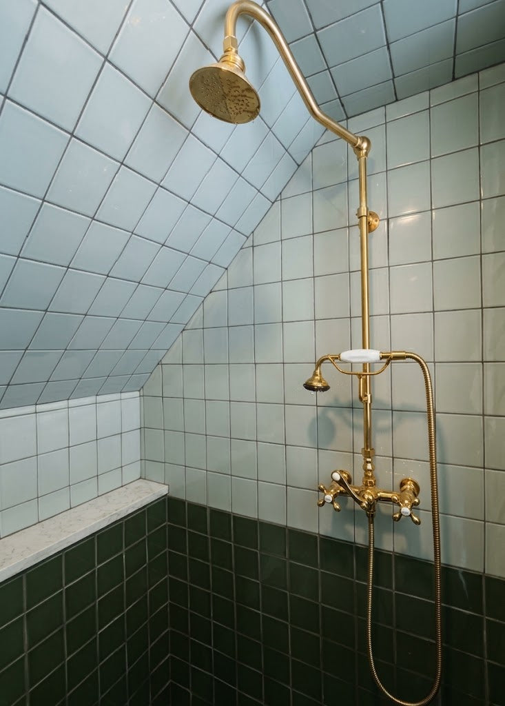 A vintage-style brass shower fixture with a round rain head, adjustable wall-mounted arm, handheld shower hose, and dual temperature controls, installed in a tiled shower corner with pale blue upper tiles and dark green lower tiles.