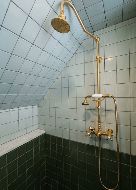 A vintage-style brass shower fixture with a round rain head, adjustable wall-mounted arm, handheld shower hose, and dual temperature controls, installed in a tiled shower corner with pale blue upper tiles and dark green lower tiles.