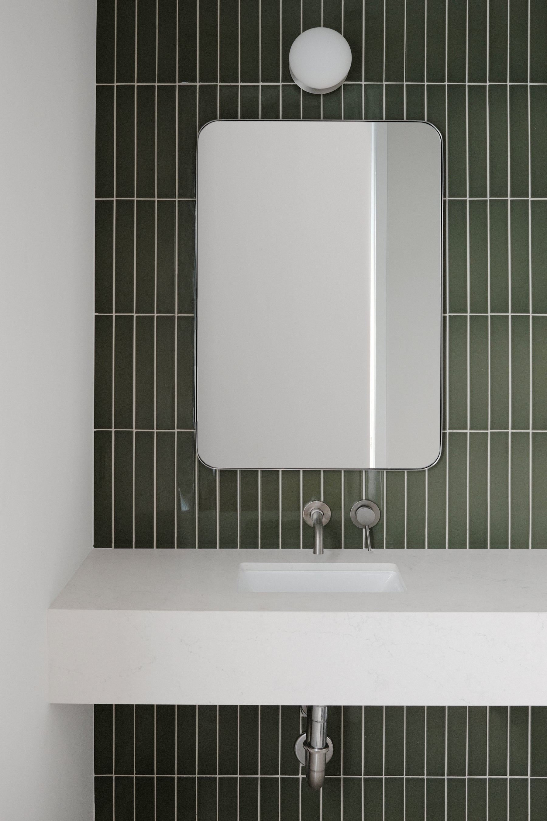 Dark green glossy solid tile by Clay Imports in a vertical pattern behind a white vanity with a mirror.