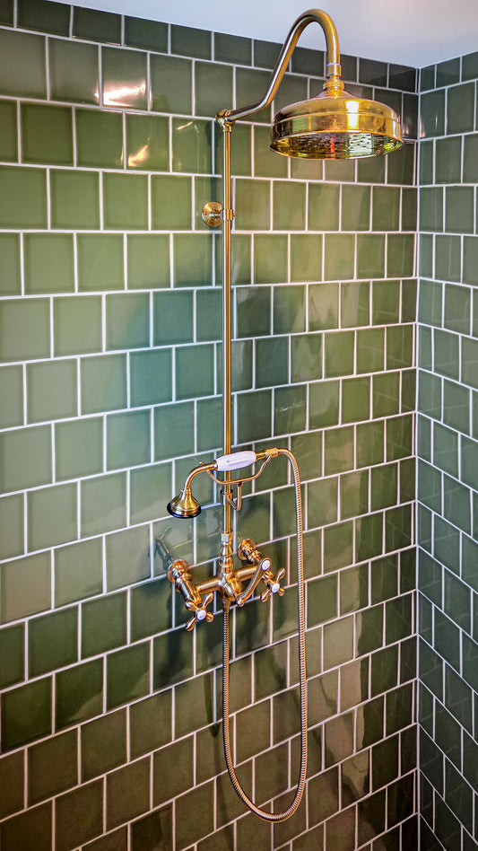 A brass shower fixture with a round rain head, adjustable hand shower, and flexible hose, mounted against a green subway tile wall.