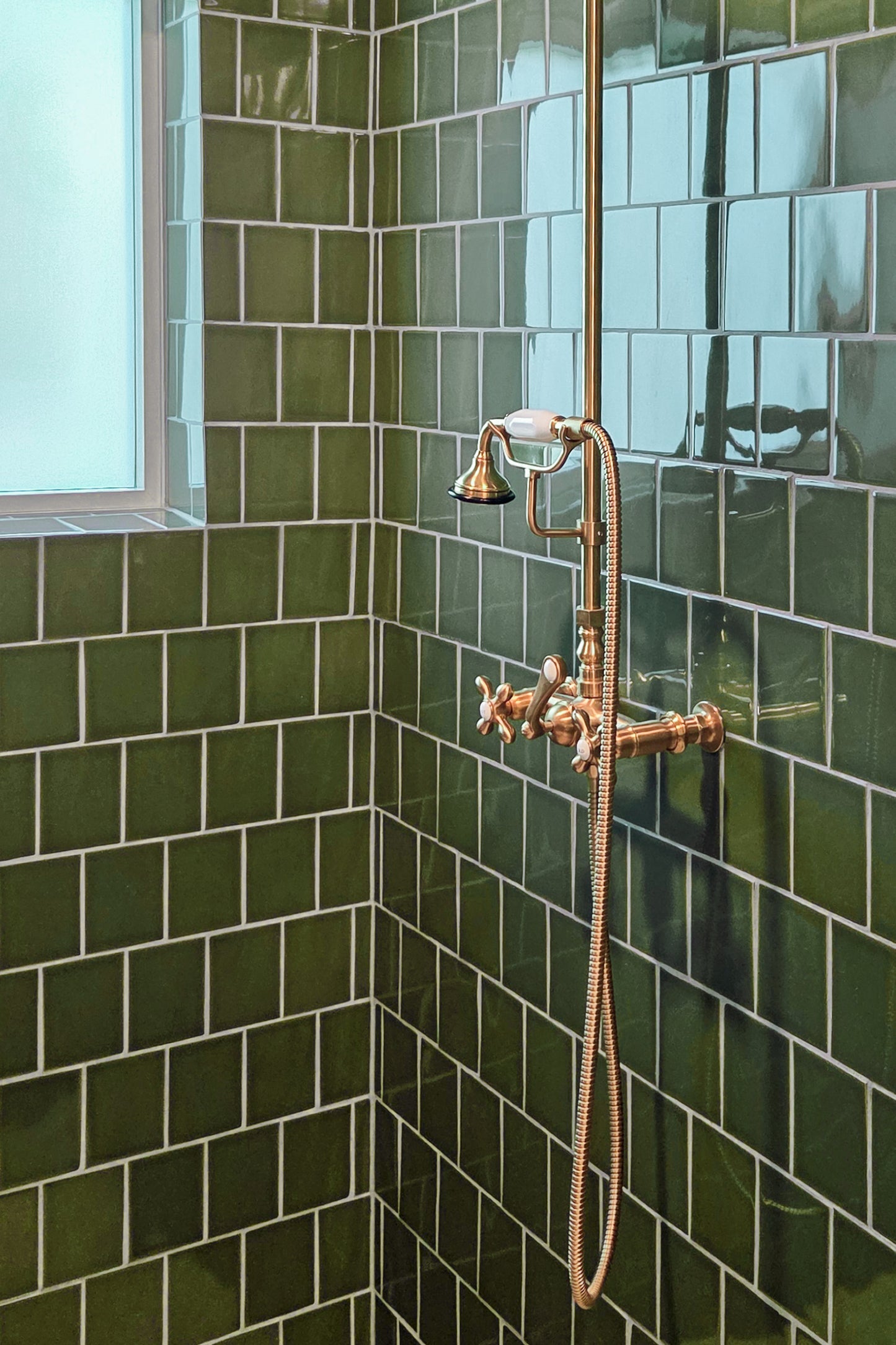 A close-up of a bathroom corner with glossy dark green subway tiles and a copper-toned wall-mounted shower fixture with a handheld shower head and flexible hose.