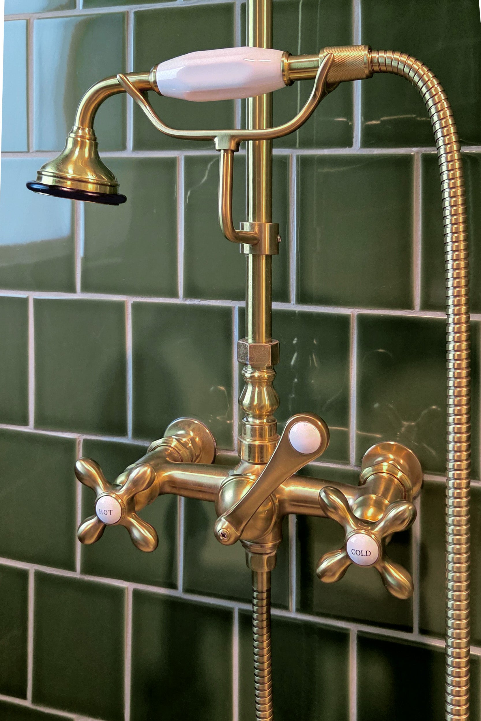 An antique brass tub filler faucet set with a handheld shower, mounted against a green ceramic tile wall.