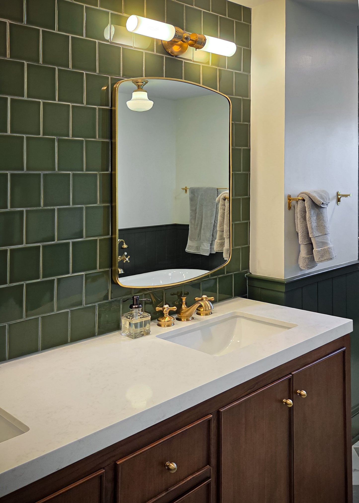 A modern bathroom vanity featuring a white countertop, dark wood cabinets, a large rectangular gold-framed mirror, and a green subway tile backsplash.