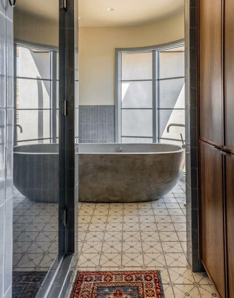 A modern freestanding concrete bathtub sits in a brightly lit bathroom with patterned tile flooring and arched windows.