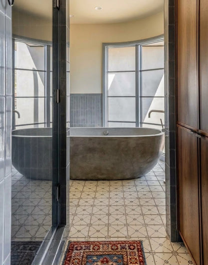 A modern freestanding concrete bathtub sits in a brightly lit bathroom with patterned tile flooring and arched windows.