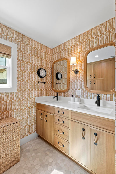 A double-sink bathroom vanity with light wood cabinets, a white countertop, and patterned wallpaper in beige and brown tones. Two wood-framed mirrors, a wall clock, and a wall-mounted sconce are visible, along with a woven basket chair and a window on the left.