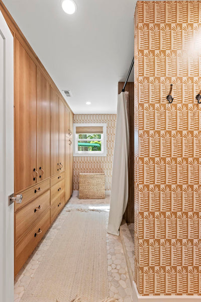 A bathroom hallway with light wood cabinetry on the left, a patterned orange and beige tiled wall on the right, a white shower curtain, and a small window at the end of the hall.
