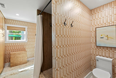 A modern bathroom interior with patterned beige and white wallpaper, a white toilet, a freestanding wicker storage bench, a window with a green-tinted shade, and a shower curtain.