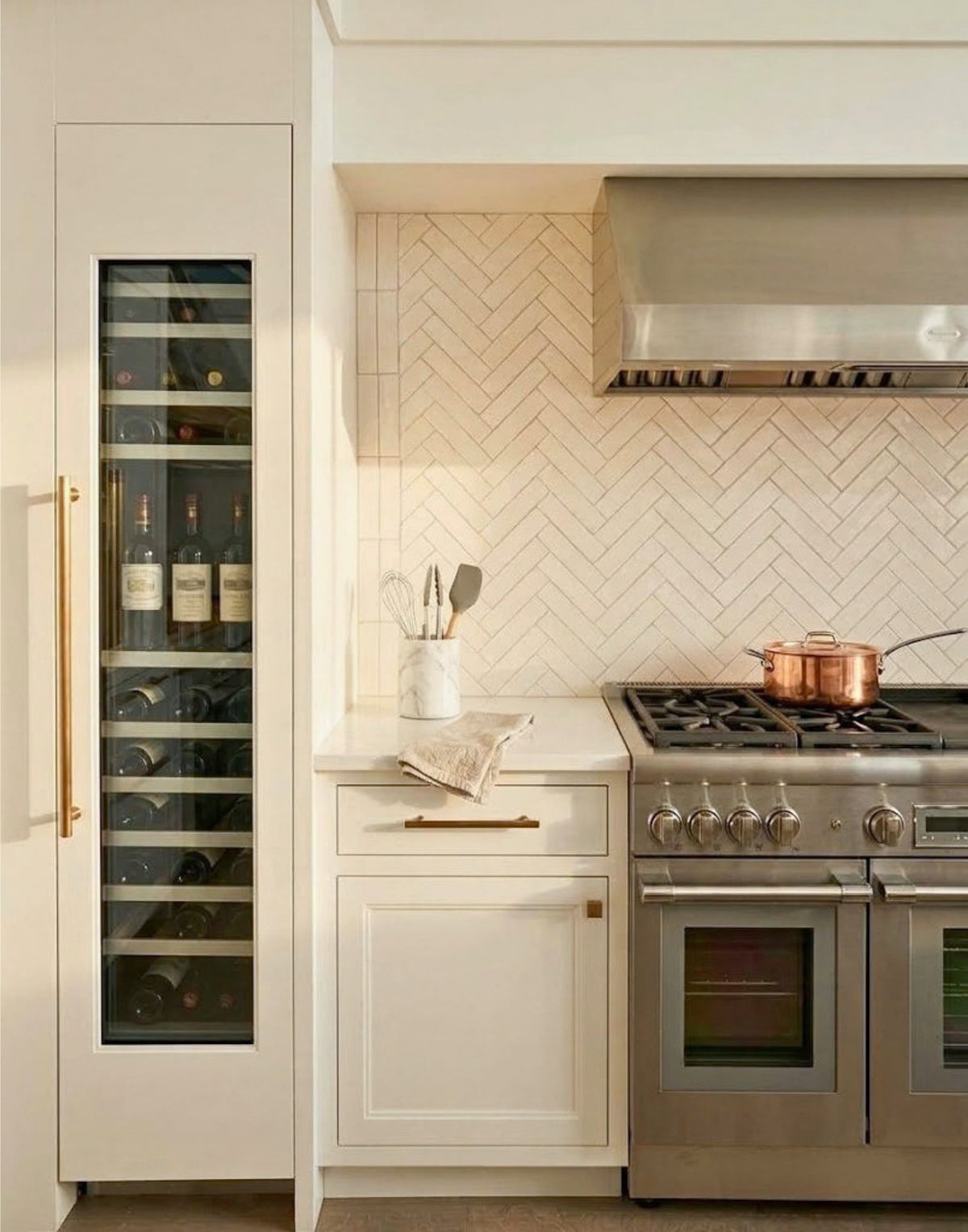 A modern kitchen features a tall built-in wine refrigerator, a stainless steel range stove with a copper pot, and elegant white herringbone tile backsplash.