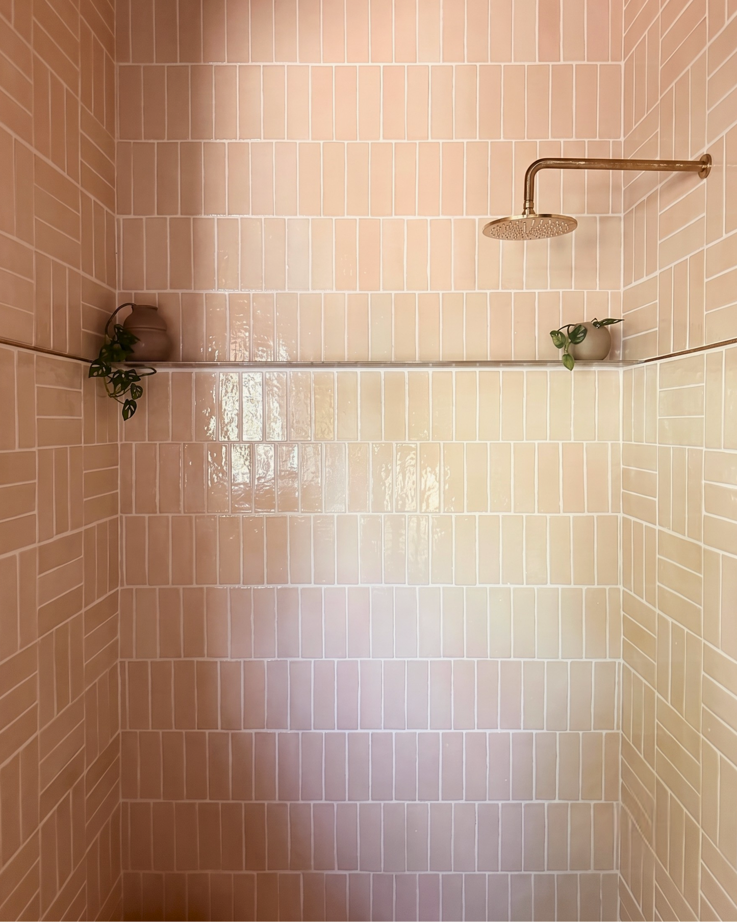 A shower wall with glossy vertical blush pink subway tiles, a round shower head mounted on the upper right, and two small potted green plants on the shelf.