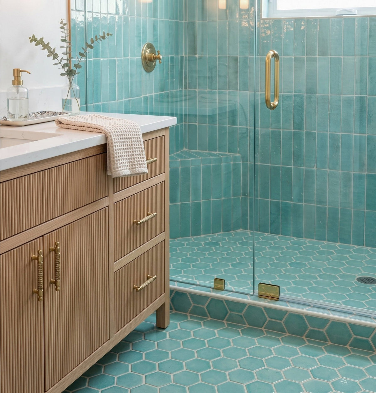 A light wood vanity with fluted drawers and brass hardware stands beside a walk-in shower featuring glossy teal zellige tile and hexagonal floor tiles.