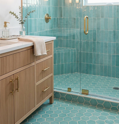 A light wood vanity with fluted drawers and brass hardware stands beside a walk-in shower featuring glossy teal zellige tile and hexagonal floor tiles.