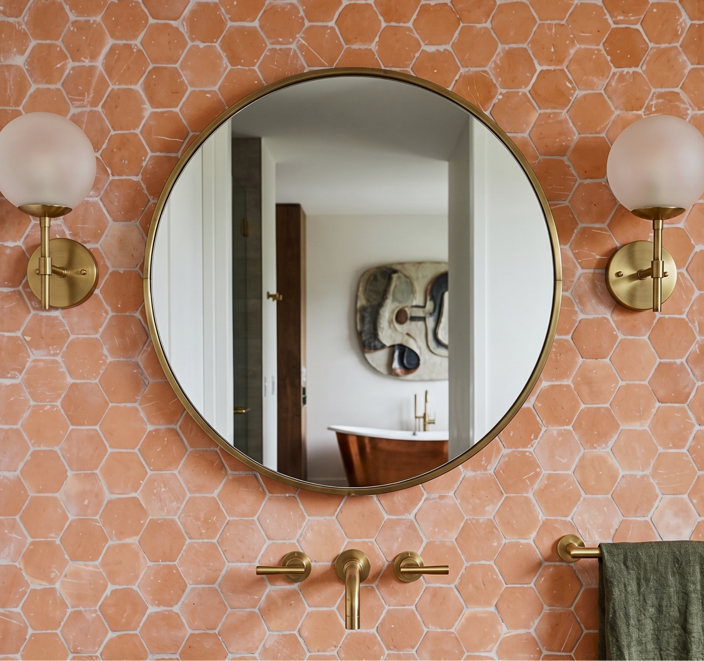 Round wall mirror with a gold-toned frame mounted on a terracotta hexagonal tile wall, flanked by two matching gold wall sconces and brass bathroom fixtures. 