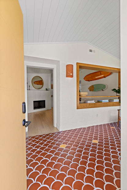 An interior room view showing a terracotta and white scallop-patterned tile floor, white paneled walls and ceiling, a built-in wood shelf with surfboards, and a doorway on the left with a yellow door frame.