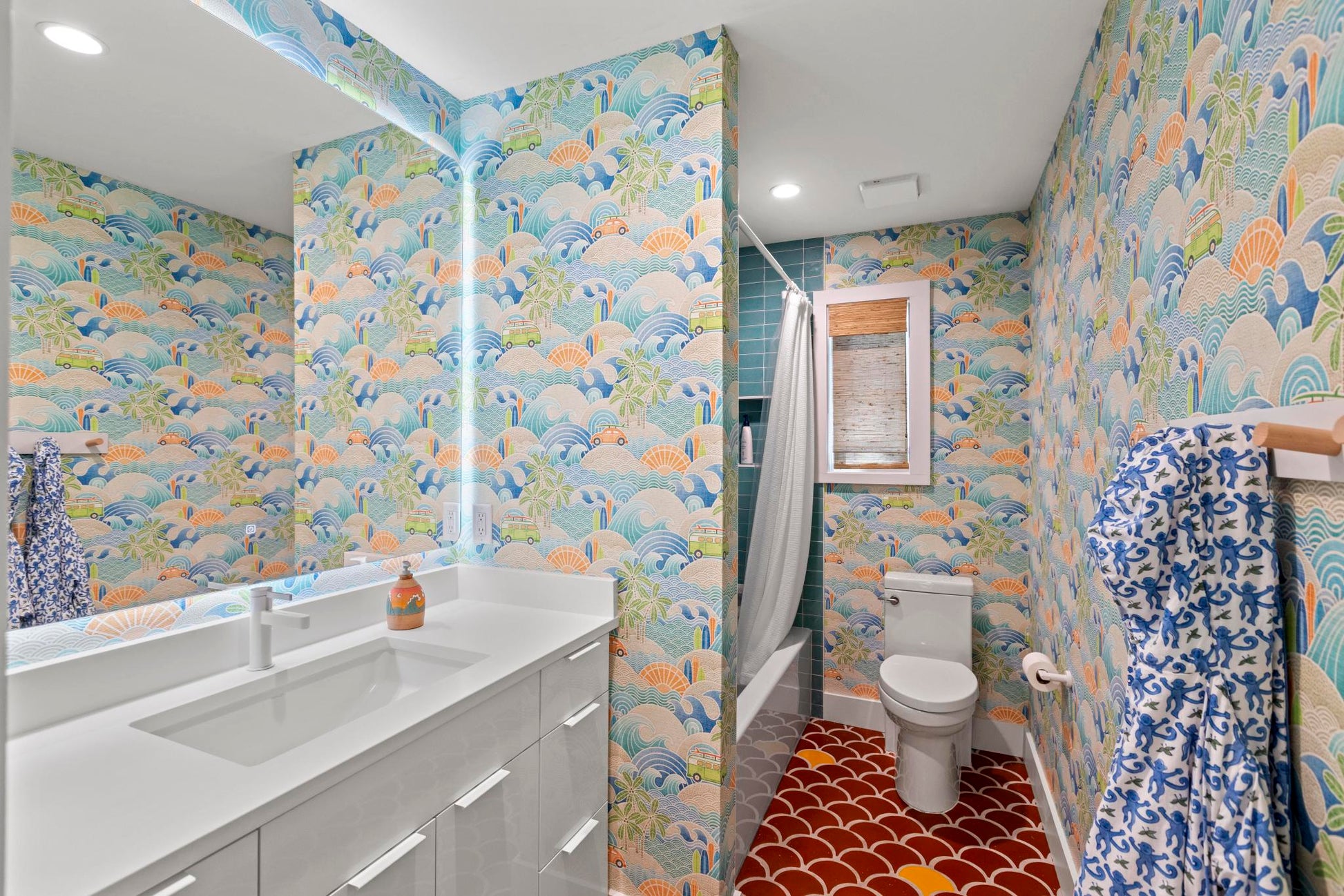 A modern bathroom featuring vibrant, multicolored tropical and wave-patterned wallpaper, a white double-sink vanity, a white toilet, and a red patterned tile floor.