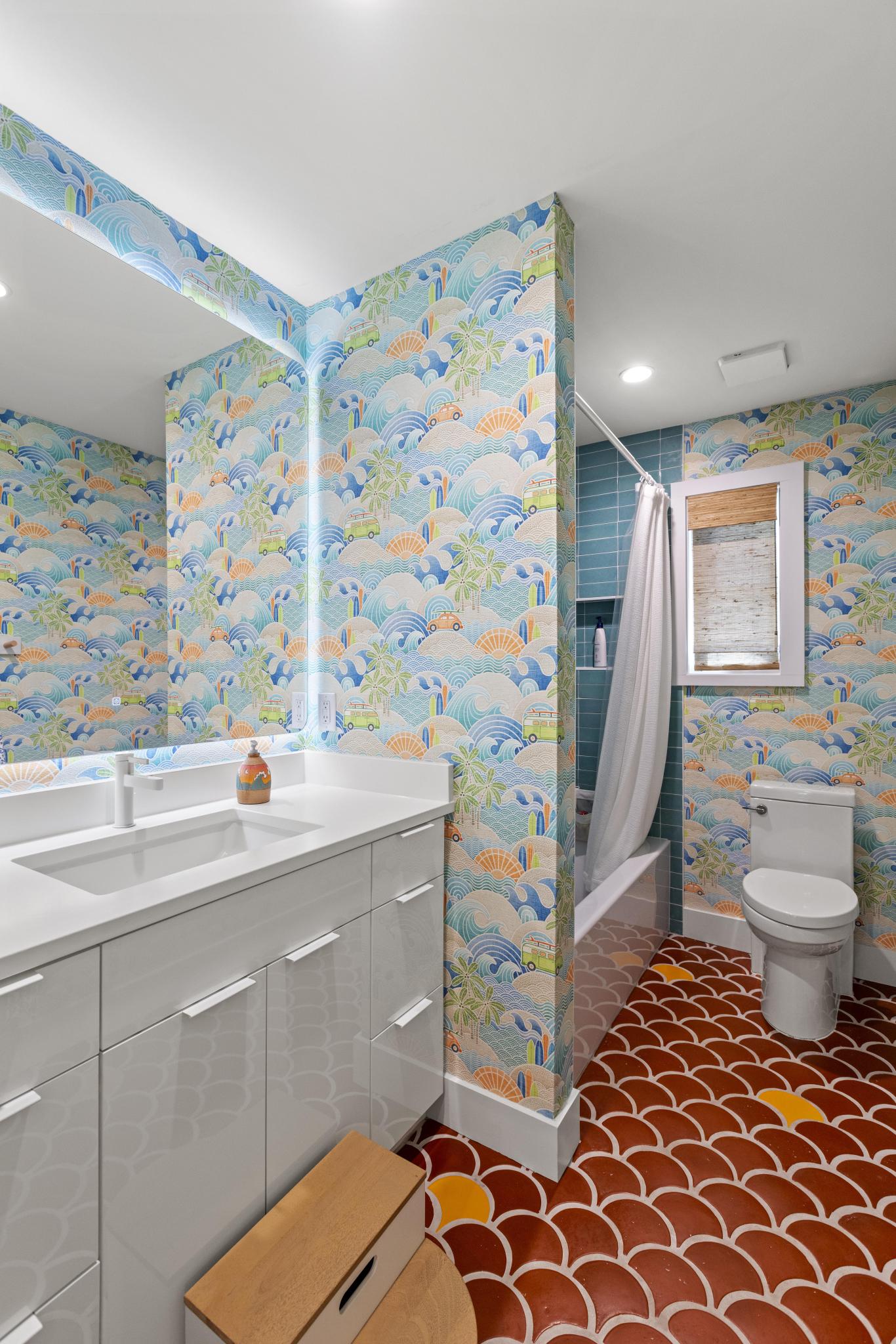 A modern bathroom interior with colorful ocean-wave pattern wallpaper, a white vanity with sink, a toilet, a shower curtain, and a red scalloped tile floor.