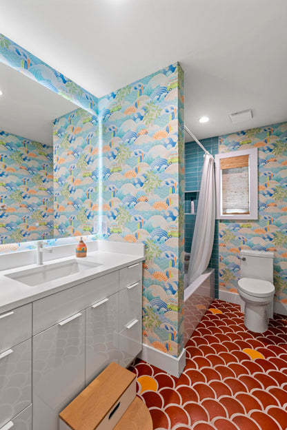 A modern bathroom interior with colorful ocean-wave pattern wallpaper, a white vanity with sink, a toilet, a shower curtain, and a red scalloped tile floor.