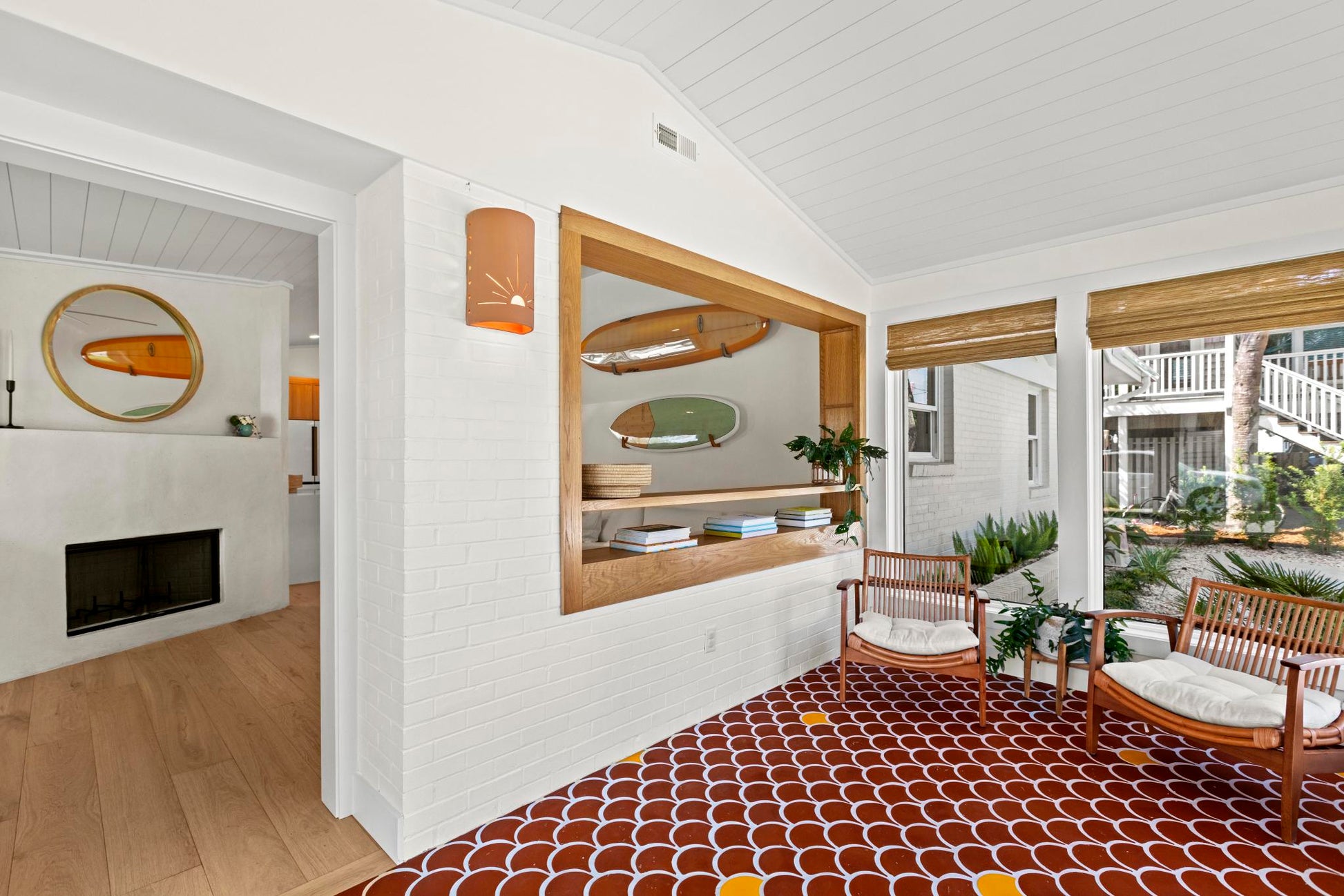 A modern interior space with white brick walls, a red and white scalloped patterned tile floor, two wooden lounge chairs with white cushions, and surfboard-shaped wall decor.