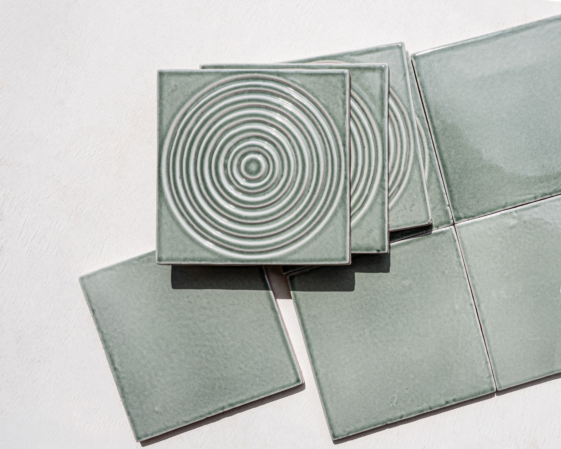 Square ceramic tiles in a muted sage green glaze, with one tile stacked on top showing a raised concentric circle pattern, photographed on a plain light background.