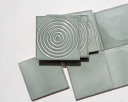 Square ceramic tiles in a muted sage green glaze, with one tile stacked on top showing a raised concentric circle pattern, photographed on a plain light background.