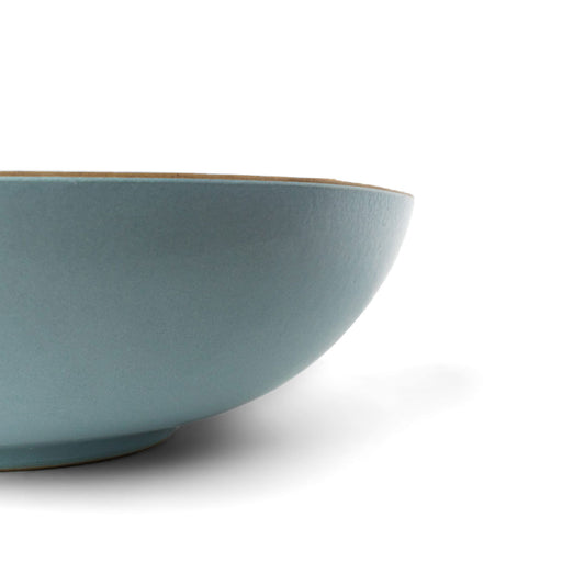 A large dusty blue matte ceramic salad bowl by Clay Imports with a natural tan rim, shown from a side angle.