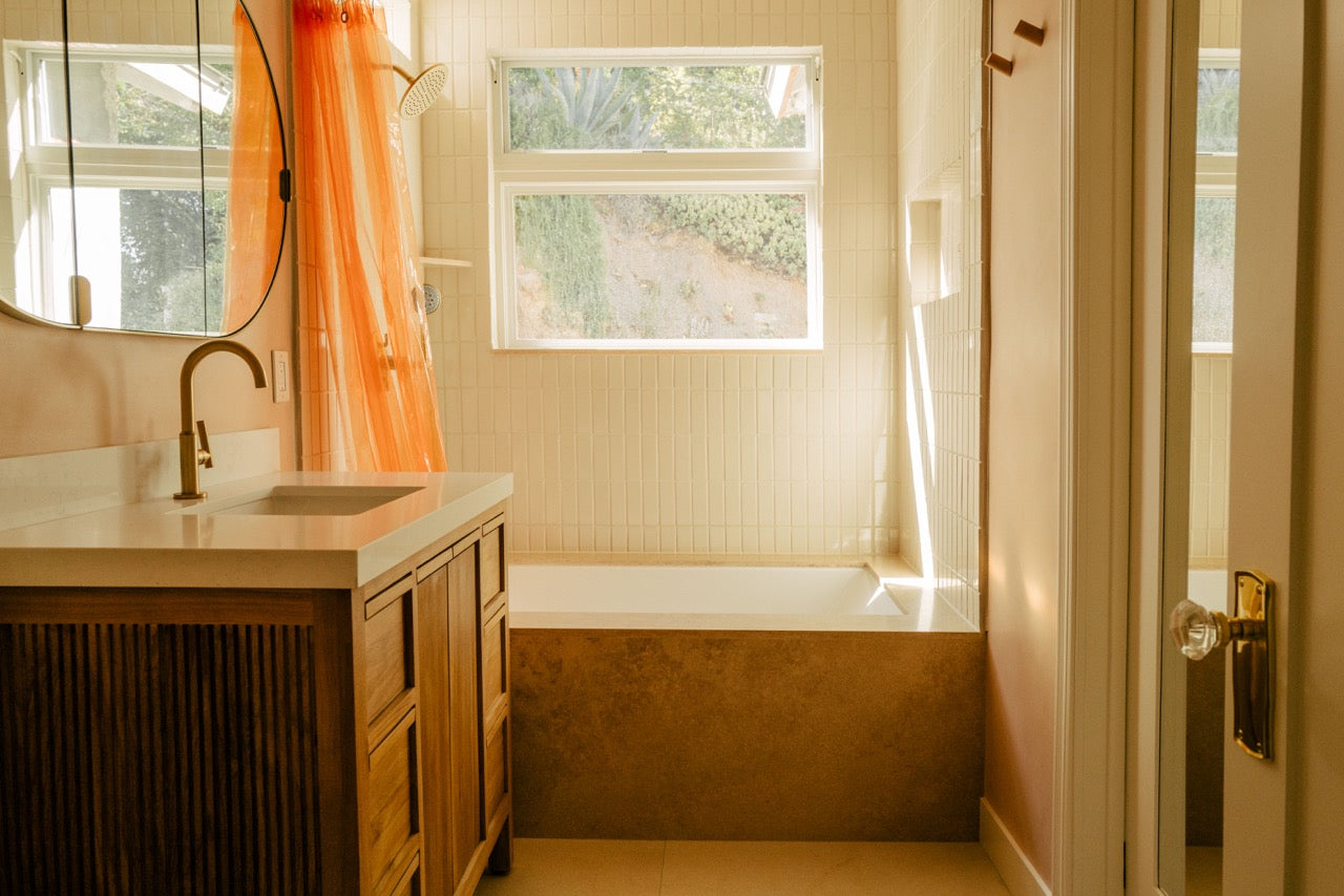 A warm, sunlit bathroom interior featuring a wooden vanity with a white countertop and sink, a white bathtub with a terracotta-colored base, a bright orange shower curtain, and a window looking out onto greenery.