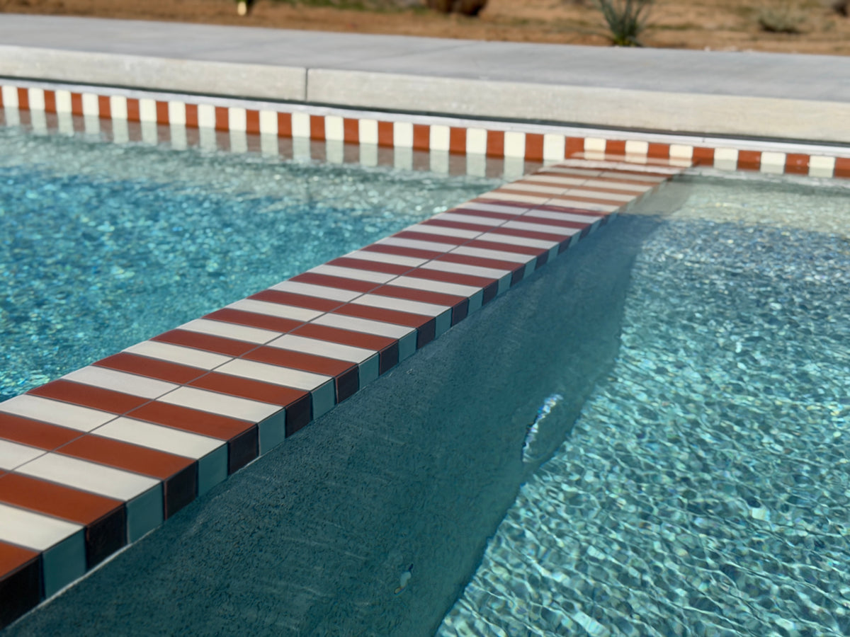 Mamey red and white striped 2x6 pool waterline tile by Clay Imports Handmade Tile, with a submerged bench and shimmering blue water.