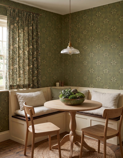 A cozy dining nook with olive green floral patterned wallpaper, a built-in wooden bench with cream cushions, a round wooden table, two wooden chairs, and a hanging glass pendant light.