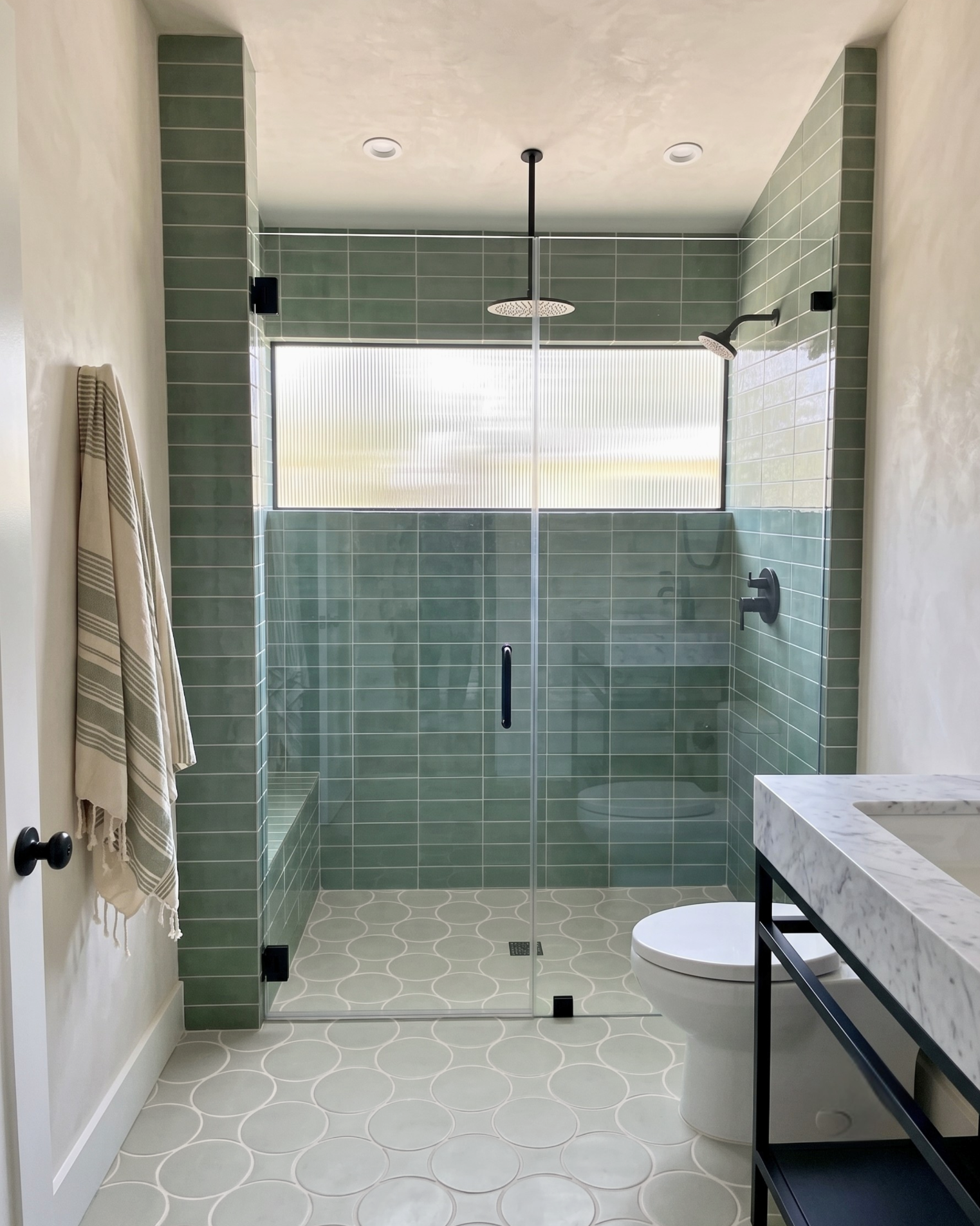A modern bathroom featuring a walk-in shower with a frosted window, sage green tile walls and floor, a white toilet, and a marble countertop with a black frame.