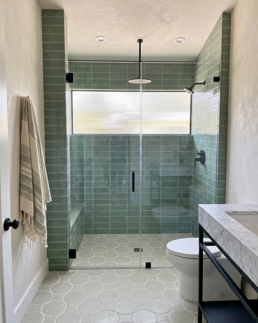 A modern bathroom featuring a walk-in shower with a frosted window, sage green tile walls and floor, a white toilet, and a marble countertop with a black frame.