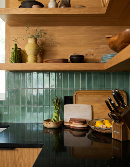 A contemporary kitchen features warm oak open shelving, glossy green zellige tile backsplash, and a sleek black countertop styled with a bunch of fresh asparagus, a bowl of lemons, and wooden cutting boards.