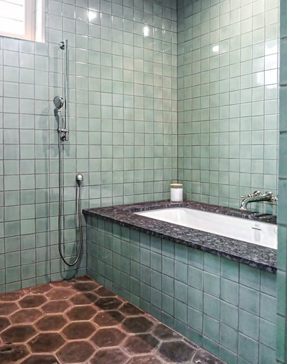 A bathroom corner with pale green square wall tiles, a built-in white bathtub with a dark stone surround, a wall-mounted shower fixture, and dark hexagonal floor tiles.