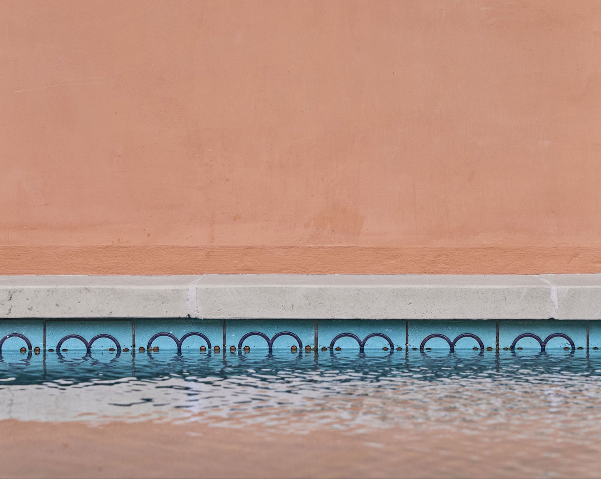 Blue-green relief pool waterline tile by Clay Imports, featuring a repeating dark blue semi-circle pattern, reflecting in rippled water against a peach wall.