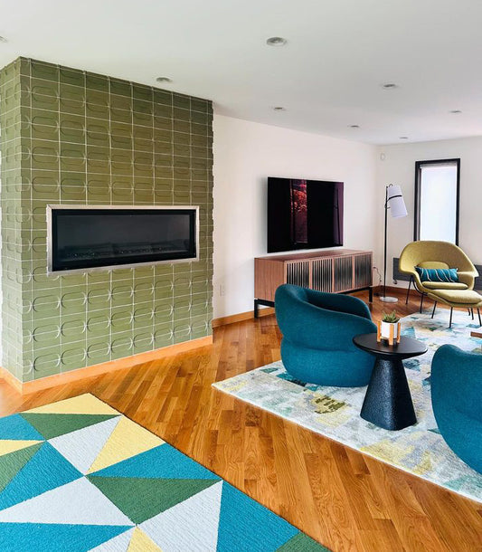 A modern living room interior with a large green tiled wall, built-in fireplace, wood flooring, teal armchairs, a patterned rug, and a black side table.