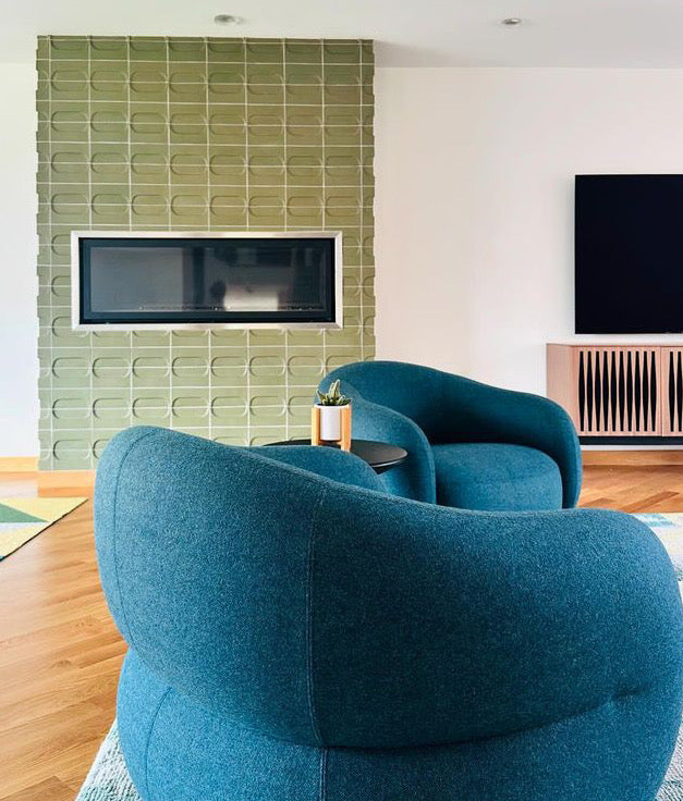 A modern living room featuring two curved teal blue armchairs in the foreground, with a textured sage green tiled wall and a built-in fireplace behind them.