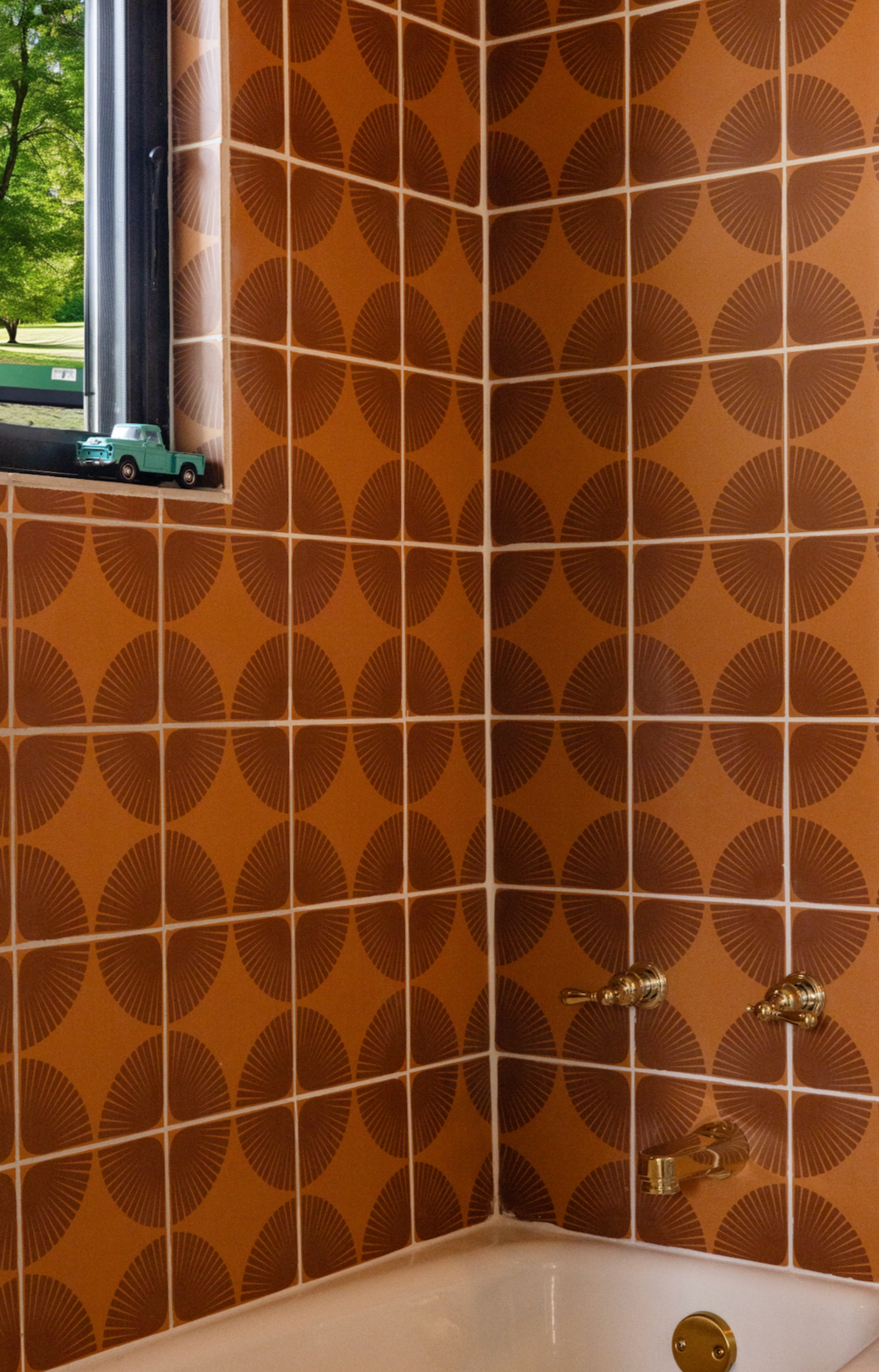 Orange-brown pattern tile by Clay Imports with dark brown petal designs, on a bathroom wall surrounding a window and white tub.