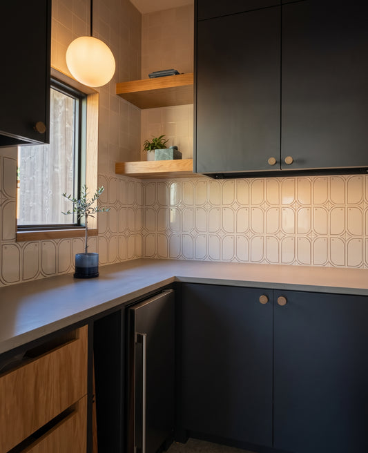 A modern kitchen corner features dark blue cabinets, light wood open shelving, patterned ceramic backsplash tiles, and a small potted plant near a bright window.