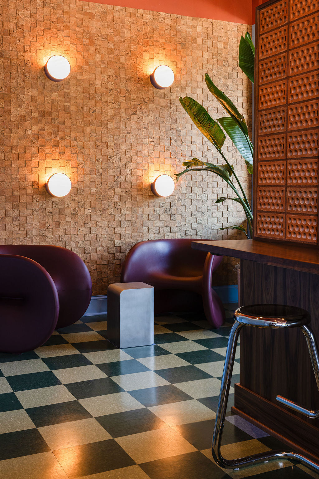 A modern interior scene with two dark purple lounge chairs, a checkered black and beige tile floor, warm wall sconces, a woven wood wall, and a wooden counter with a metal stool.
