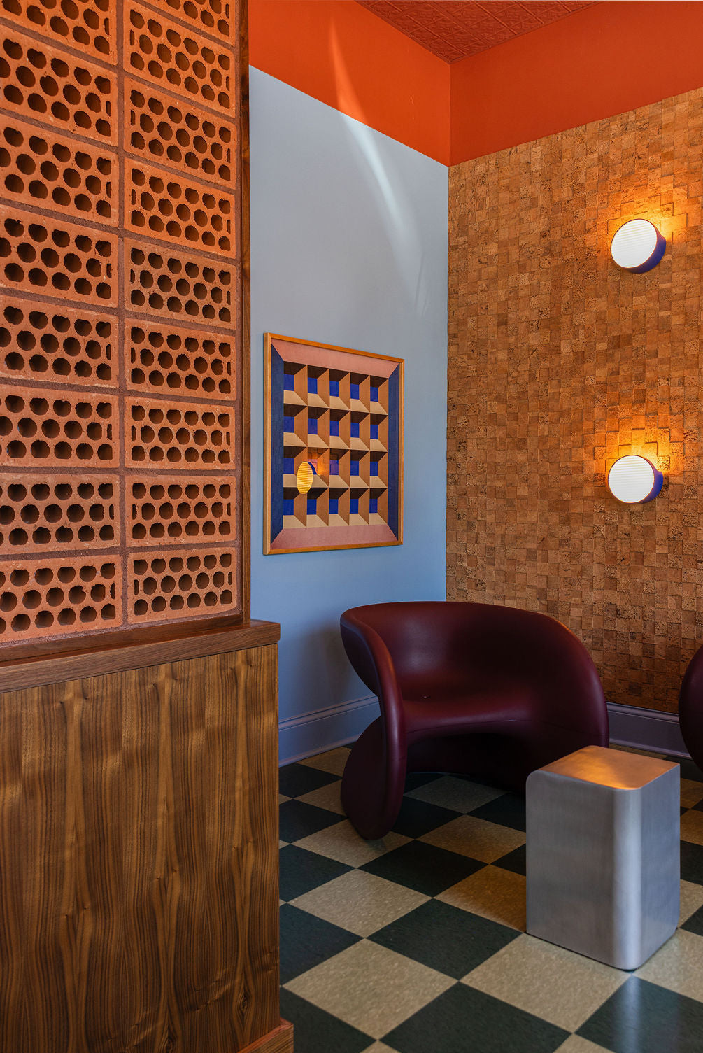 A modern interior corner with a sculptural burgundy chair, a small metallic side table, checkered black and beige flooring, a wood-paneled wall, and a patterned orange and blue accent wall.