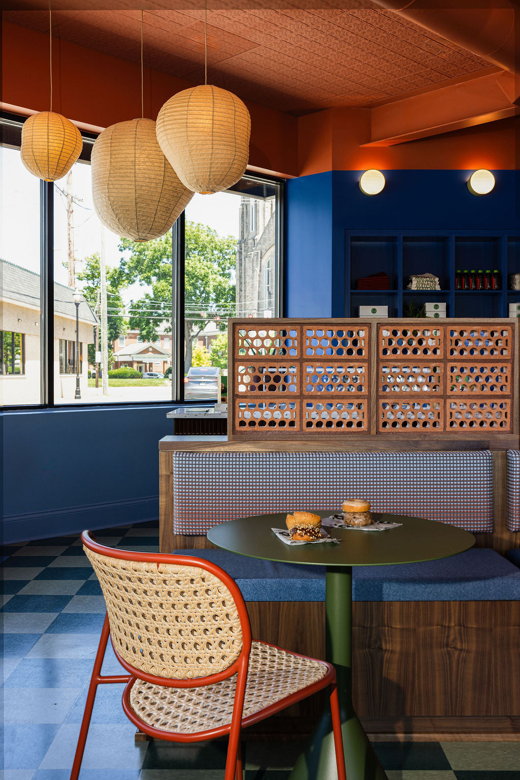 A cozy cafe interior featuring a small round green table, a woven cane chair with a red frame, a blue cushioned booth, and a wooden lattice partition. Large pendant paper lanterns hang from a textured orange-brown ceiling, and a window on the left shows a street view.