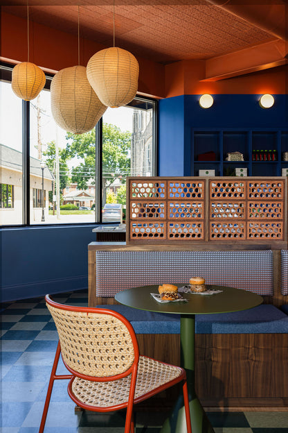 A cozy cafe interior featuring a small round green table, a woven cane chair with a red frame, a blue cushioned booth, and a wooden lattice partition. Large pendant paper lanterns hang from a textured orange-brown ceiling, and a window on the left shows a street view.