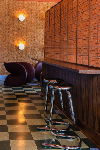 A modern interior scene with a dark wood counter and two chrome bar stools in the foreground, a checkered floor, and a textured wall with two round wall lights and curved purple seating in the background.