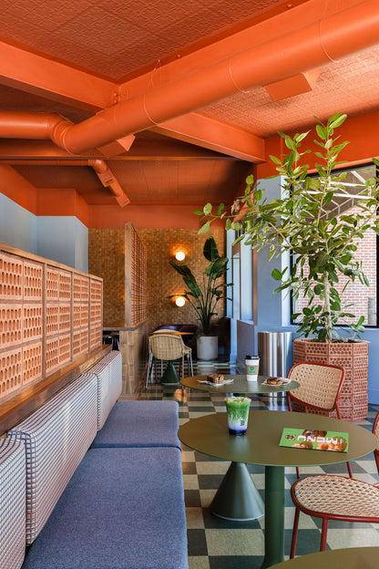 A cozy restaurant or cafe interior with orange ceiling and exposed pipes, blue upholstered booth seating, round green tables, woven chairs, checkered flooring, and large potted plants.