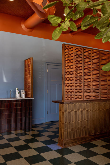 An interior architectural scene with a wooden slatted counter and matching decorative wall panels, a blue-gray wall, a black-and-white checkered floor, and hanging green leaves overhead.
