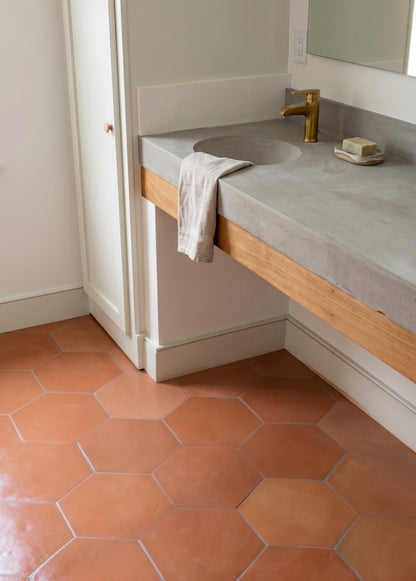 A modern concrete bathroom vanity with a brass faucet rests above terracotta hexagonal floor tiles.
