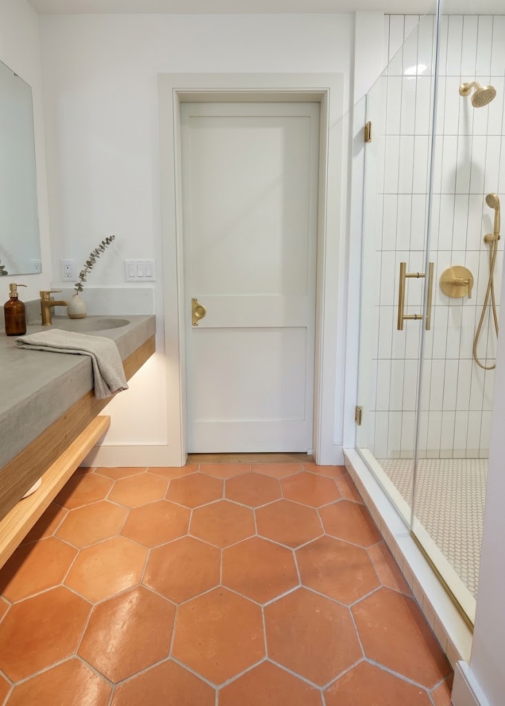 A modern bathroom features terracotta hexagon floor tiles contrasting with a floating concrete vanity and a glass shower enclosure with brushed brass fixtures.