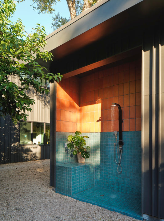 An outdoor shower area built into a modern building, with orange and teal tiled walls and floor, a dark metal shower fixture, gravel ground, and greenery around it.