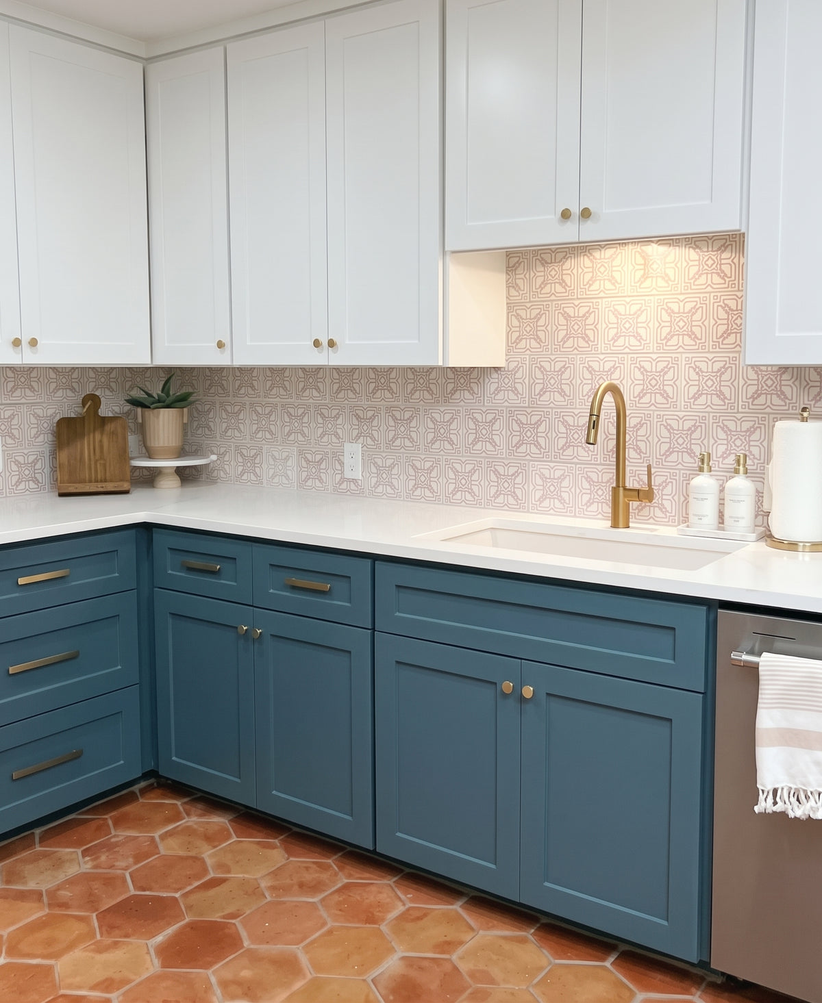 A modern kitchen features white upper cabinets, deep blue lower cabinets, a brass faucet over a white farmhouse sink, and pink patterned tile backsplash.