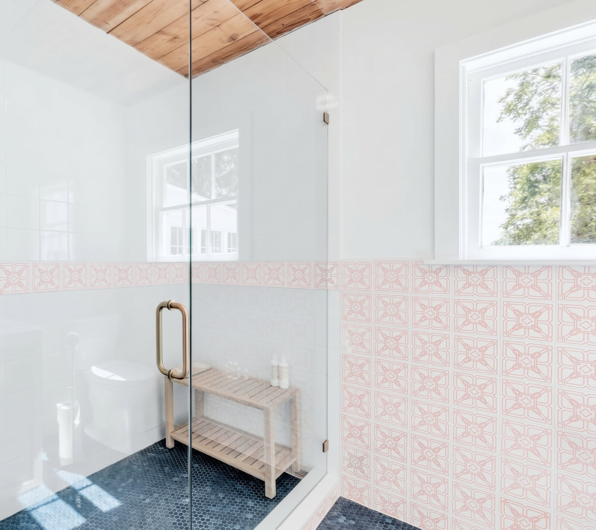 A bright modern bathroom features a glass-enclosed shower with a wooden bench, dark penny tile flooring, and walls decorated with pink and white patterned tile.
