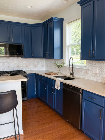 A modern kitchen featuring dark blue cabinets, light beige countertops, a white patterned tile backsplash, and warm hardwood floors. A black bar stool is visible in the foreground.